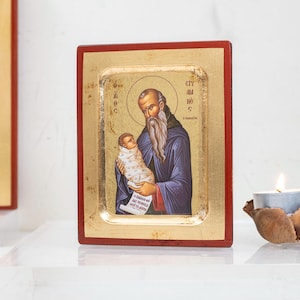 May include: A religious icon depicting a man with a long white beard holding a baby. The icon has a gold background and a red border. A second icon is partially visible on the left. A lit candle and flowers are in the background.