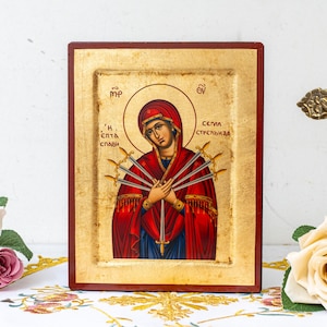 May include: A religious icon depicting a woman in a red robe and head covering, pierced by seven swords. The icon is framed in a dark wood border with gold accents. A gold crucifix and roses are in the background.