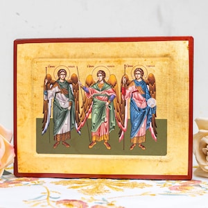 May include: A framed religious icon featuring three figures with halos, wings, and robes in shades of green, pink, and blue. The figures hold spears and a sphere. The frame is red with a gold background.
