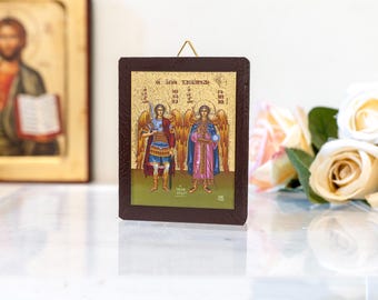 May include: A framed religious icon depicting two angels with golden wings and halos. The angels are standing on a green background with a gold border. The text "OI AΓIOI TAXIAΡΧΕΣ" is written in Greek at the top of the icon.