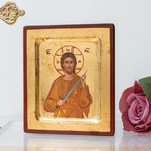 May include: A religious icon depicting a figure with a halo, holding a sword. The icon is set in a gold-toned frame with a dark red border. A gold cross and roses are in the background.