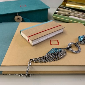 Puede incluir: Dos diarios marrones con tapas azul verdoso, uno pequeño y otro grande, están sobre una superficie blanca. Un collar plateado con un colgante circular y dos alas con piedras de imitación turquesa está sobre el diario más grande.