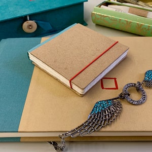 Puede incluir: Una colección de artículos de papelería que incluye un cuaderno pequeño con una banda elástica roja, un diario más grande con una cubierta turquesa y un colgante plateado en forma de ala con detalles turquesas. También se ve una caja turquesa y una caja verde y dorada.