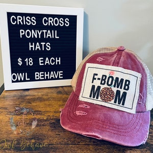 May include: A distressed burgundy ponytail hat with a tan mesh back. The hat features a patch with the text "F-BOMB MOM" and a leopard print design. A sign displays "CRISS CROSS PONYTAIL HATS" and "OWL BEHAVE".