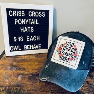 May include: A distressed black and gray ponytail hat with a mesh back. The hat features a square patch with the text "Ugh, There's People Here" surrounded by floral designs. A white framed sign reads "Criss Cross Ponytail Hats".