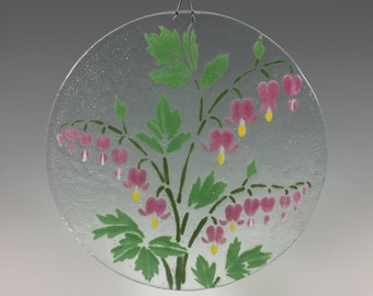 May include: A clear, round glass hanging decoration featuring a painted design of a bleeding heart flower. The flowers are pink with yellow accents, and the leaves are green. A silver wire is attached for hanging.