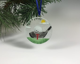 May include: A circular glass Christmas ornament with a golf theme. It depicts a golf ball on a tee, a golf club, a green field, a yellow sun, and a blue ribbon. The background is light grey.