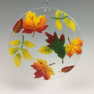 May include: A clear, round glass ornament featuring colorful autumn leaves in shades of red, orange, yellow, and green. The leaves are painted on the glass, and a metal wire is attached for hanging. The ornament is a decorative item.