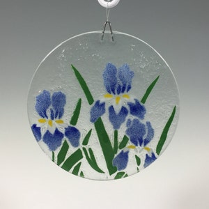 May include: A round, clear glass suncatcher featuring a design of blue iris flowers with yellow centers and green leaves. The suncatcher is suspended by a silver wire and hook, perfect for a window or wall decoration.