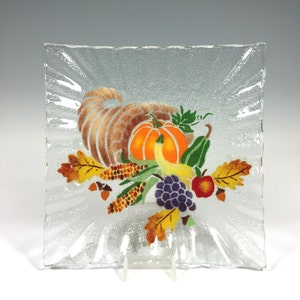Puede incluir: Plato cuadrado de vidrio transparente con superficie texturizada y diseño temático de Acción de Gracias. El diseño presenta una cornucopia rebosante de calabazas, uvas, maíz y hojas de otoño en tonos naranja, amarillo y marrón.