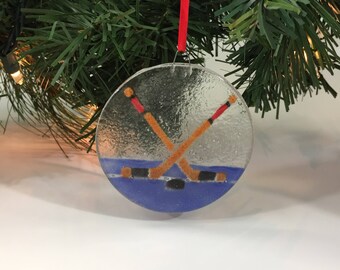 Può includere: Una decorazione natalizia rotonda in vetro trasparente con un tema di hockey. L'ornamento presenta due mazze da hockey incrociate, un disco e uno sfondo blu e argento. Un nastro rosso è attaccato per appenderlo.
