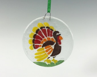 May include: A round, clear glass ornament featuring a colorful turkey design. The turkey has a brown body, red and yellow tail feathers, and is set against a white background. A green ribbon is attached for hanging. A festive Thanksgiving decoration.