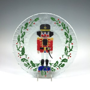 May include: A clear glass plate featuring a colorful depiction of a nutcracker soldier. The plate is decorated with a green holly wreath and red berries around the edge. The nutcracker is dressed in a red coat, black boots, and a tall black hat. A festive holiday decoration.