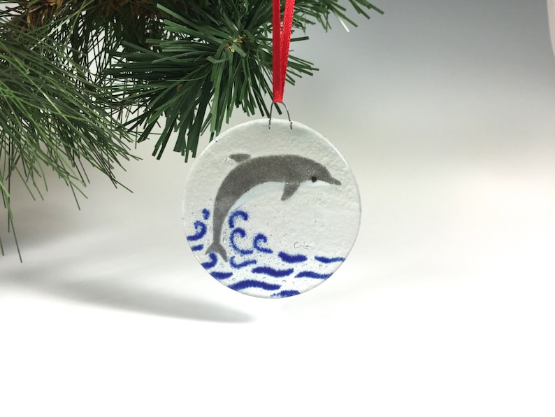 May include: Circular glass Christmas decoration depicting a grey dolphin jumping from blue waves. The ornament hangs from a red ribbon, set against a backdrop of evergreen foliage.