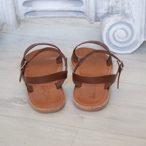 K&ouml;nnte beinhalten: Ein Paar braune Ledersandalen mit hellbrauner Sohle und d&uuml;nnen Riemen, die an der Kn&ouml;chelseite geschnallt werden. Die Sandalen sind auf einer hellen Holzoberfl&auml;che ausgestellt. Klassisches Design.