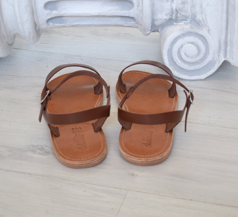 K&ouml;nnte beinhalten: Ein Paar braune Ledersandalen mit hellbrauner Sohle und d&uuml;nnen Riemen, die an der Kn&ouml;chelseite geschnallt werden. Die Sandalen sind auf einer hellen Holzoberfl&auml;che ausgestellt. Klassisches Design.
