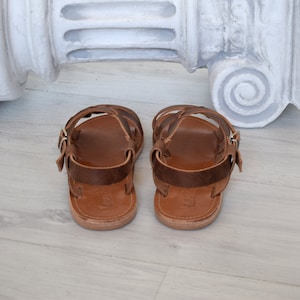 May include: A pair of brown leather sandals with multiple straps and a buckle closure. The sandals have a light brown sole and are designed for comfort and style. The footwear is displayed on a light-colored wooden surface.