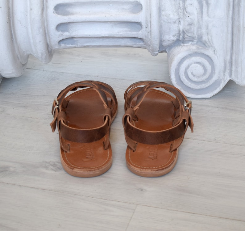 May include: A pair of brown leather sandals with multiple straps and a buckle closure. The sandals have a light brown sole and are designed for comfort and style. The footwear is displayed on a light-colored wooden surface.