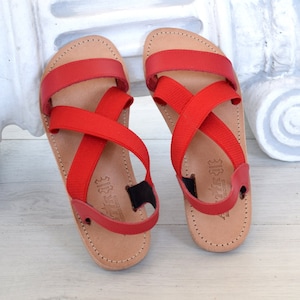 May include: Red leather sandals with criss-cross straps. The straps are made of a red elastic material.