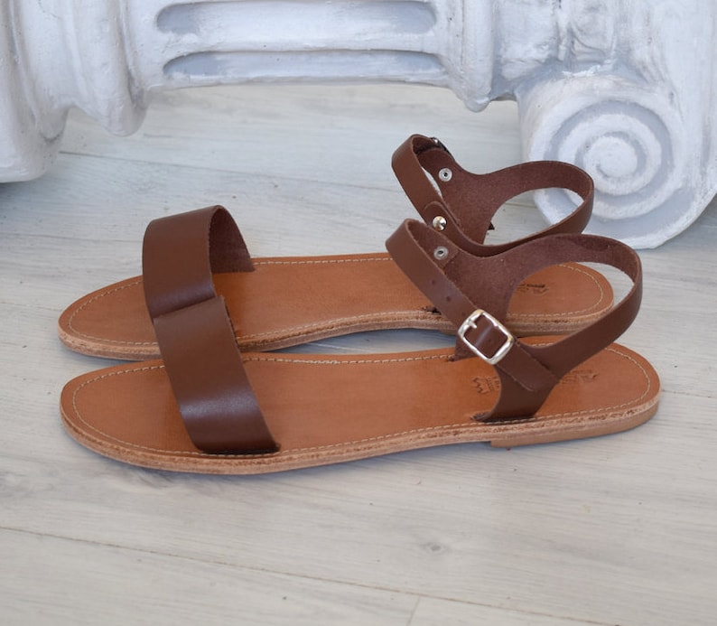K&ouml;nnte beinhalten: Braune Ledersandalen mit schlichtem Design. Die Sandalen haben einen einzelnen Riemen &uuml;ber den Zehen und einen Kn&ouml;chelriemen mit einer silbernen Schnalle. Das Fu&szlig;bett ist hellbraun.