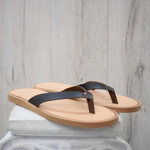 May include: A pair of leather flip-flops with dark brown straps and light brown soles. The sandals are displayed on a white surface against a light wood-grain background. The flip-flops are a neutral colour.