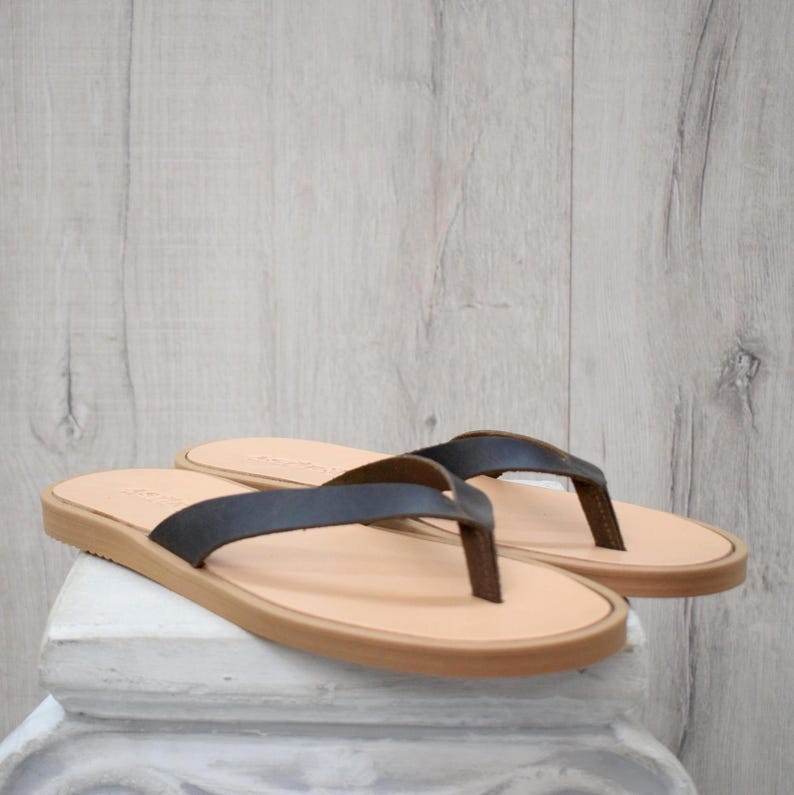 May include: A pair of leather flip-flops with dark brown straps and light brown soles. The sandals are displayed on a white surface against a light wood-grain background. The flip-flops are a neutral colour.