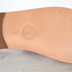 May include: A light brown leather sandal sole with a textured brown heel. The sole has a natural, smooth finish with a circular embossed logo and the number 38. The sandal sole is designed for footwear.