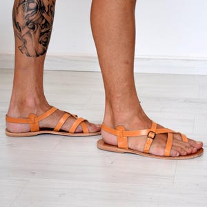 Op de afbeelding: Oranje leren sandalen met meerdere bandjes en een gespsluiting. De sandalen hebben een platte zool en zijn ontworpen voor een comfortabele pasvorm. Het schoeisel heeft een warme oranje kleur.