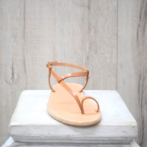 May include: A tan leather sandal featuring a thin strap design with a toe loop and an adjustable ankle strap with a small metal buckle. The flat sole has visible stitching along the edge, providing a simple and functional footwear construction.