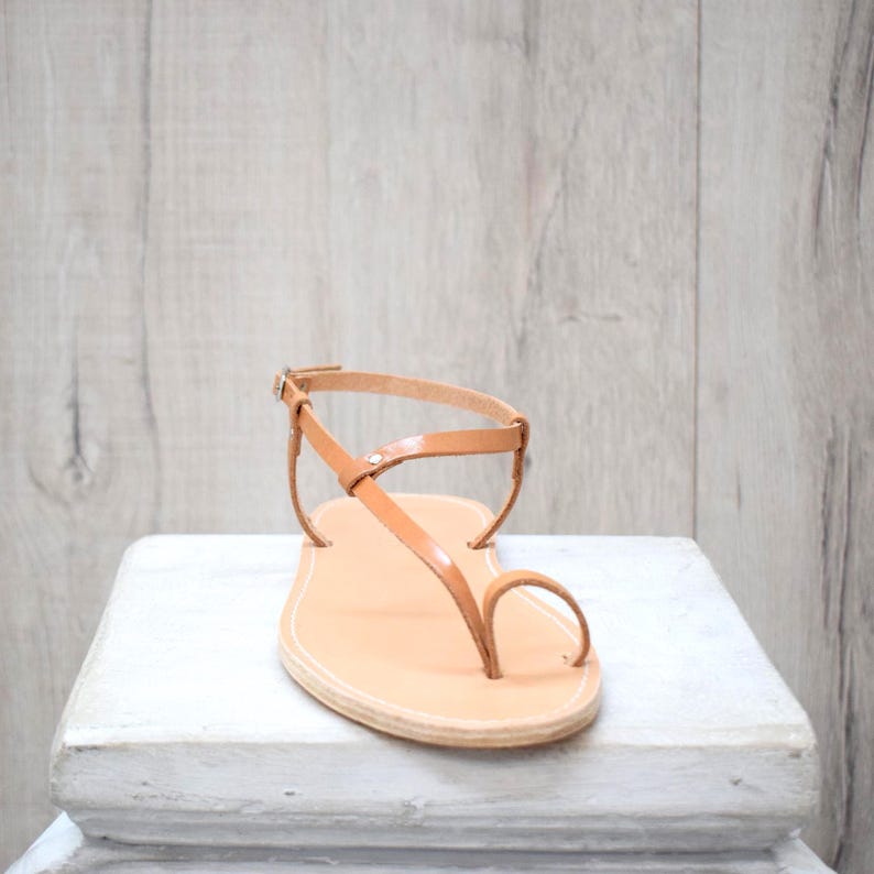 May include: A tan leather sandal featuring a thin strap design with a toe loop and an adjustable ankle strap with a small metal buckle. The flat sole has visible stitching along the edge, providing a simple and functional footwear construction.