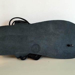 May include: The bottom of a black leather sandal with a simple design. The sandal has a single strap that wraps around the ankle and a small hole on the side.