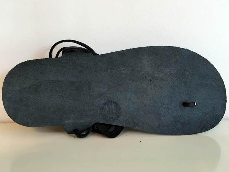 May include: The bottom of a black leather sandal with a simple design. The sandal has a single strap that wraps around the ankle and a small hole on the side.