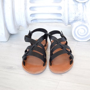 May include: A pair of black leather sandals with a crisscross strap design. The sandals have brown soles and adjustable ankle straps with metal buckles. The footwear is displayed on a light-colored wooden surface.