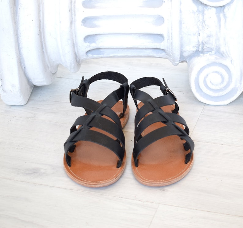 May include: A pair of black leather sandals with a crisscross strap design. The sandals have brown soles and adjustable ankle straps with metal buckles. The footwear is displayed on a light-colored wooden surface.