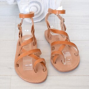 Op de afbeelding: Bruine leren sandalen met meerdere banden die over de voet en enkel kruisen. De sandalen hebben een gespsluiting aan de enkel.