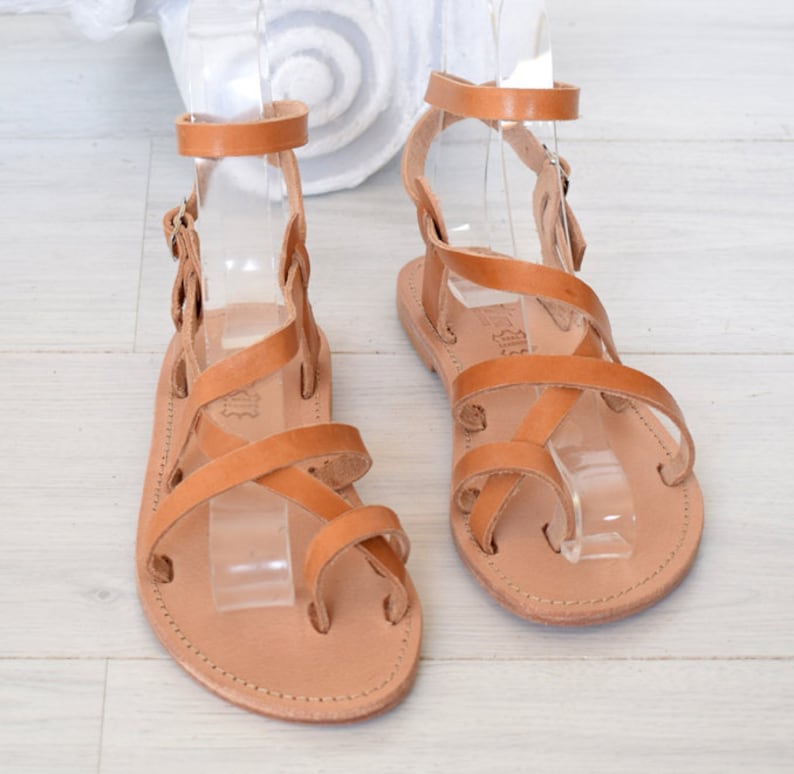 Op de afbeelding: Bruine leren sandalen met meerdere banden die over de voet en enkel kruisen. De sandalen hebben een gespsluiting aan de enkel.