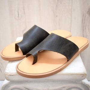 May include: A pair of dark brown leather toe-ring sandals featuring a wide strap across the top of the foot and a smooth, light tan footbed. The sandals have a flat sole and a simple, open design that secures the big toe with a leather loop.