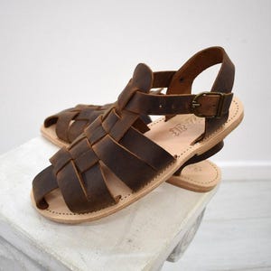 May include: Brown leather sandals with a woven design. The sandals have a strap around the ankle and a buckle closure. The soles are a light tan color. The sandals are sitting on a white surface.
