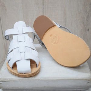 May include: White leather sandals with a woven design and a light brown sole. One sandal is upright, while the other is lying on its side, showcasing the sole. The sandals are size 38.