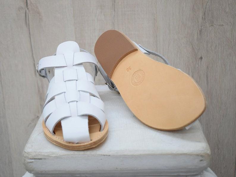 May include: White leather sandals with a woven design and a light brown sole. One sandal is upright, while the other is lying on its side, showcasing the sole. The sandals are size 38.