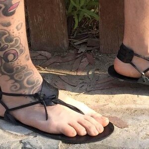 May include: A pair of black leather sandals with adjustable straps and a simple design. The sandals are worn on a person's feet with a tattoo on their leg.