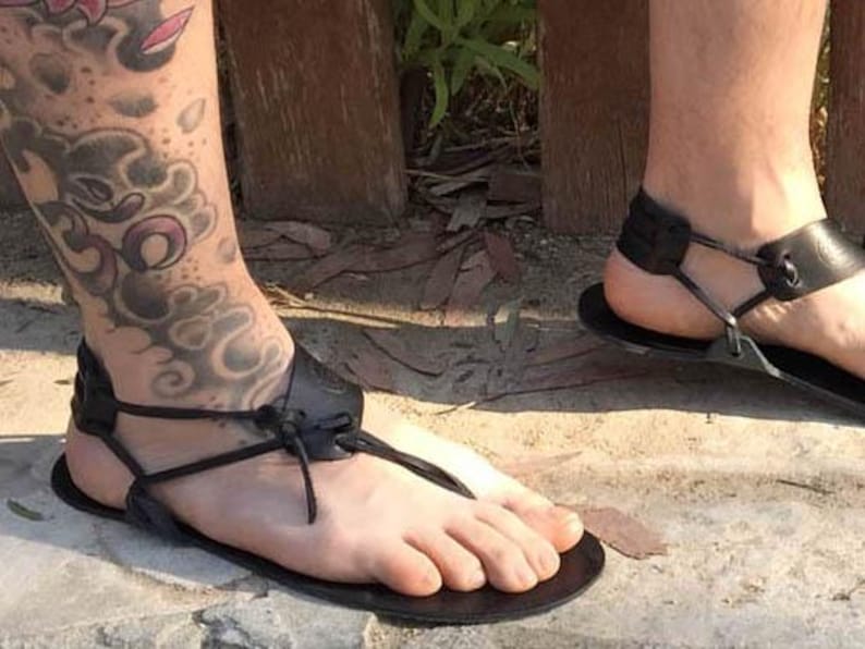 May include: A pair of black leather sandals with adjustable straps and a simple design. The sandals are worn on a person's feet with a tattoo on their leg.