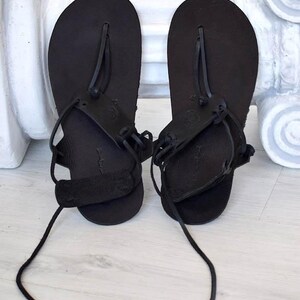 May include: A pair of black leather sandals with laces that tie up the legs. The sandals have a simple design with a single strap across the toe.