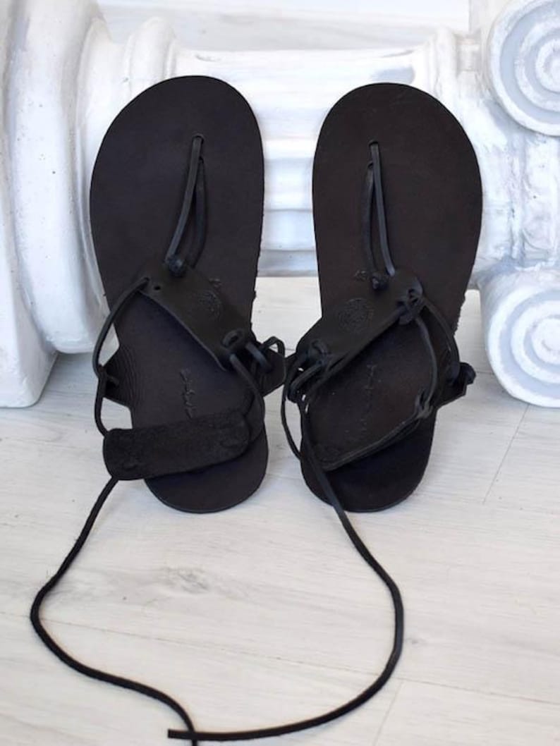 May include: A pair of black leather sandals with laces that tie up the legs. The sandals have a simple design with a single strap across the toe.