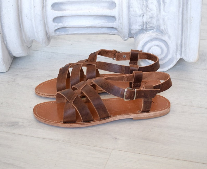 May include: A pair of brown leather sandals with multiple straps and adjustable buckles. The sandals have a flat sole and are designed for warm weather wear. The sandals are displayed on a light-colored surface.
