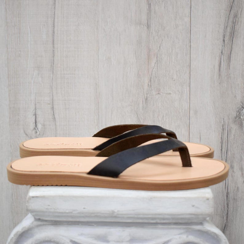May include: A pair of black leather flip-flops with a light brown sole and straps. The sandals are displayed on a white pedestal against a light wood-grain background. The flip-flops are a classic style.