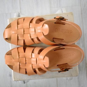 May include: A pair of light brown leather sandals with woven straps. The sandals have a buckle closure and are displayed on a light-colored surface. The sandals are a neutral color and suitable for warm weather.
