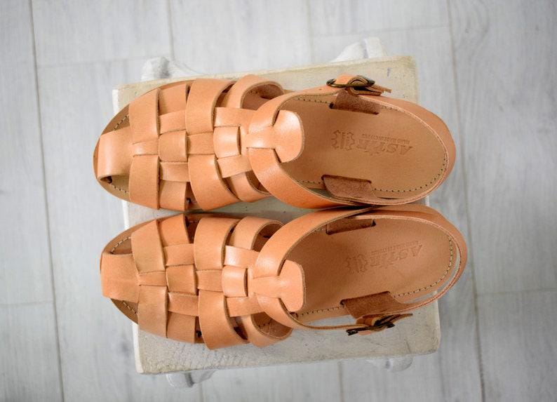 May include: A pair of light brown leather sandals with woven straps. The sandals have a buckle closure and are displayed on a light-colored surface. The sandals are a neutral color and suitable for warm weather.