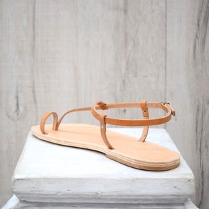 May include: A tan leather flat sandal featuring a toe loop design, a thin ankle strap with a small metal buckle, and a flat sole. The sandal has a minimalist construction with visible stitching along the edges of the straps and the footbed.