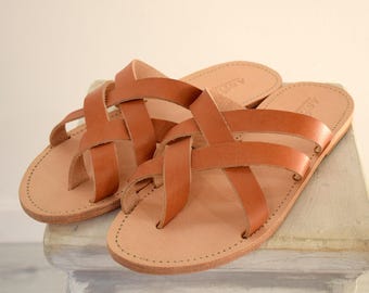 May include: A pair of tan leather sandals with a crisscross strap design. The sandals are displayed on a light-coloured surface, highlighting their simple and elegant design. The sandals are a neutral colour, suitable for various outfits.